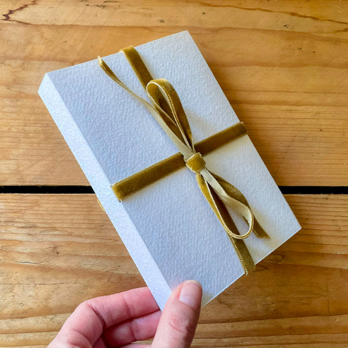 Bookbinder holds corner of Cotton Rag Sketchbook with cover and Olive Green velvet ribbon closure lies against warm, wooden surface