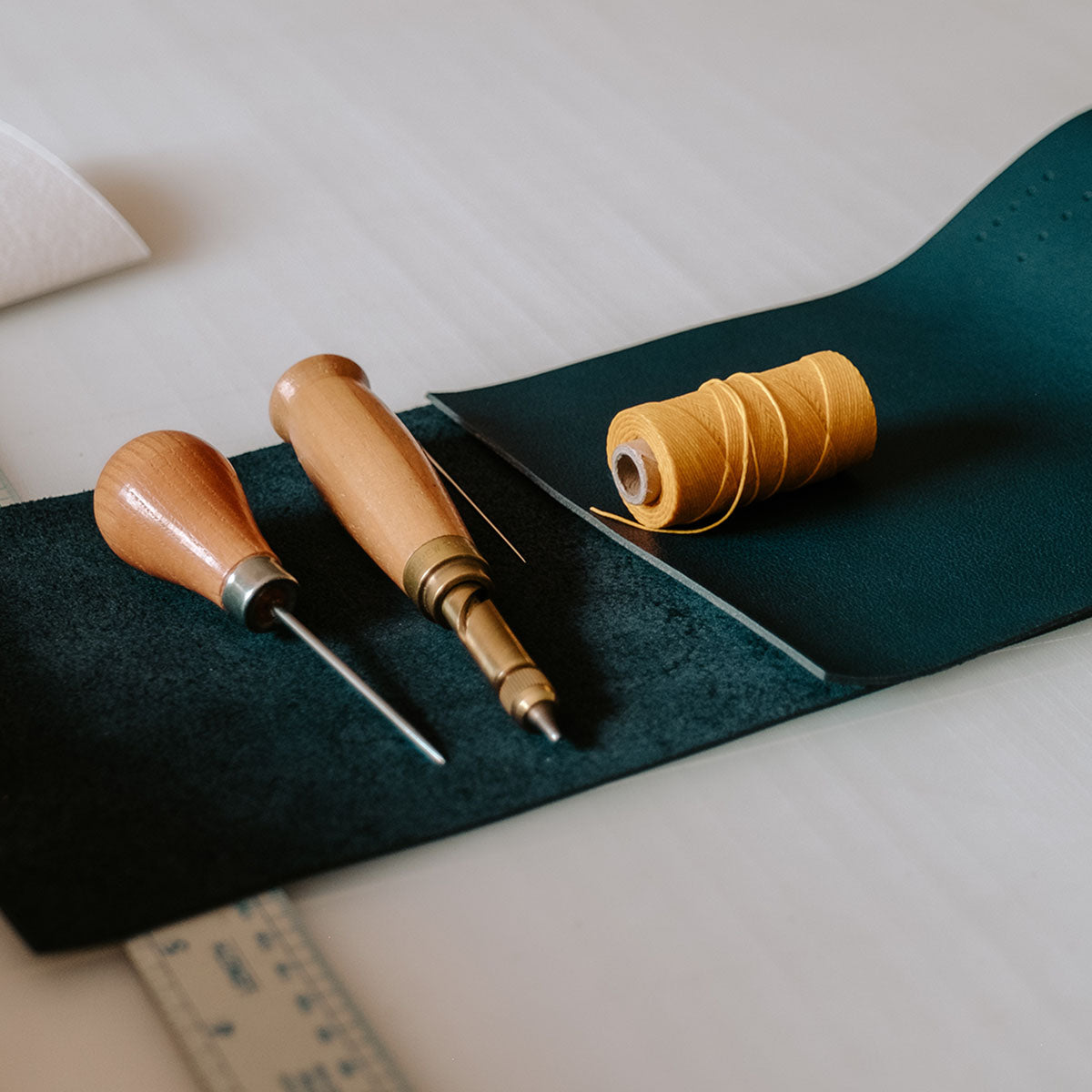 Bookbinding tools (awl, Japanese screw punch) and materials (waxed linen thread, navy leather) sit on white surface.