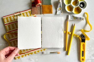 Artist holds corner of A6 Cotton Rag Sketchbook, its lay flat pages of handmade paper sit with wet and dry art materials including gouache paint, pencils and patchwork.  