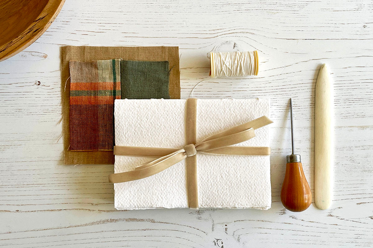 A handmade Cotton Rag Sketchbook tied with Natural Velvet Ribbon sits next to bookbinding tools and textile samples.