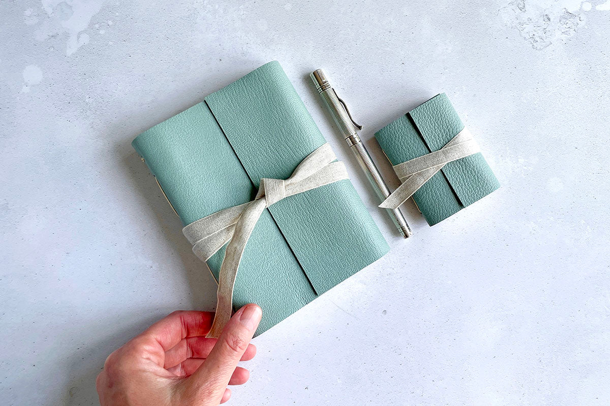 An A6 Leather Sketchbook with exposed spine stitching is held by bookbinder against a mottled grey background. Next to it sits a Mini Journal in the same colours and materials. Inbetween them lies a traditional silver fountain pen.