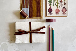 Cotton Rag Sketchbook with handmade paper and a brown velvet ribbon tied in a bow around its centre sits amongst art and scrapbooking materials including patchwork, pencils, washi tape and a postcard.