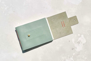 A softcover watercolour sketchbook with stud closure, bound in Duck Egg leather with Coral stitching, lies on a mottled grey surface, its closing flap open.