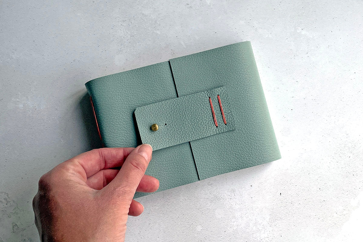 Watercolour Sketchbook bound in Duck Egg leather at Small A6 Landscape sits on blue linen cloth with watercolour palette and brush.