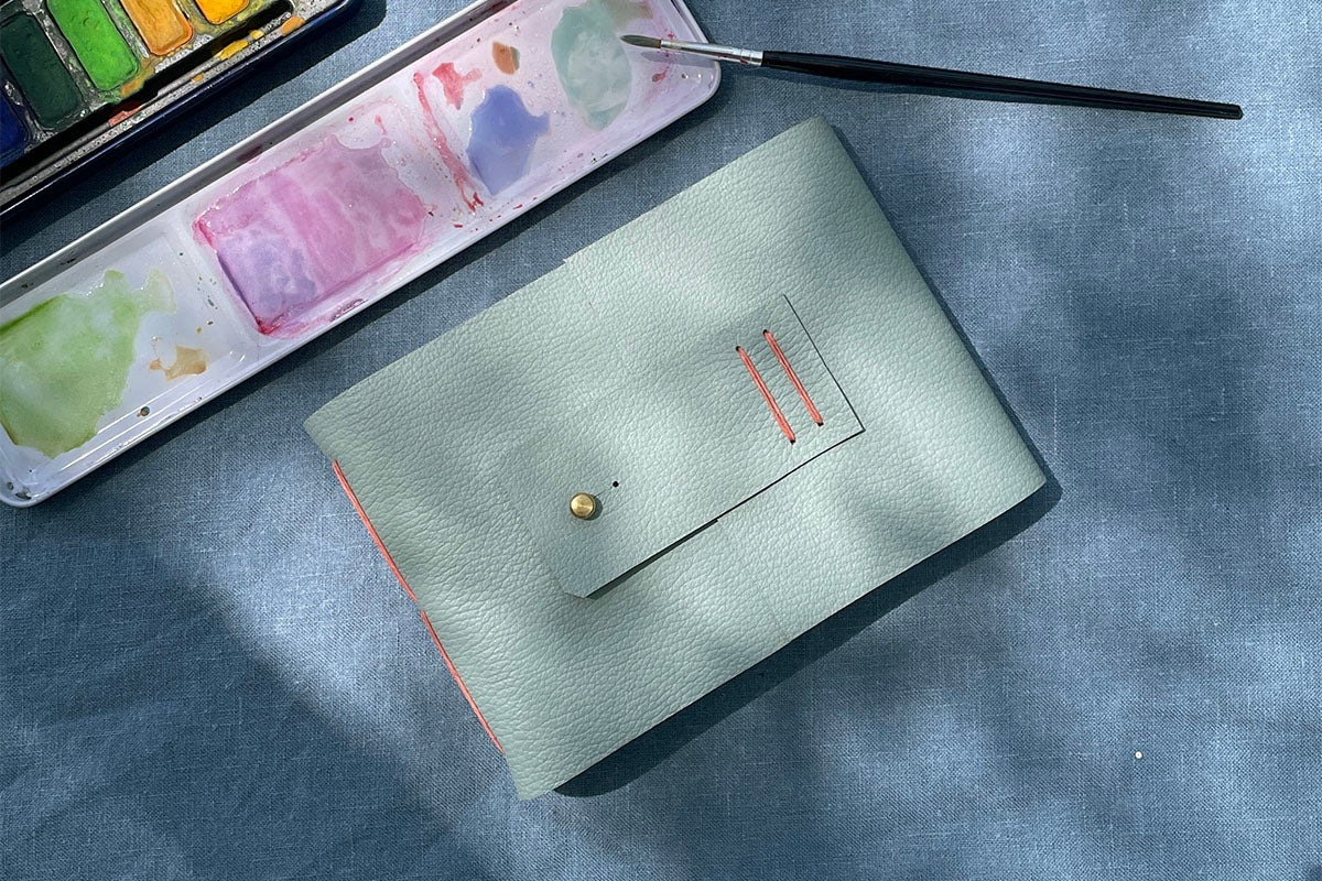 Watercolour Sketchbook bound in Duck Egg leather at Small A6 Landscape sits on blue linen cloth with watercolour palette and brush.
