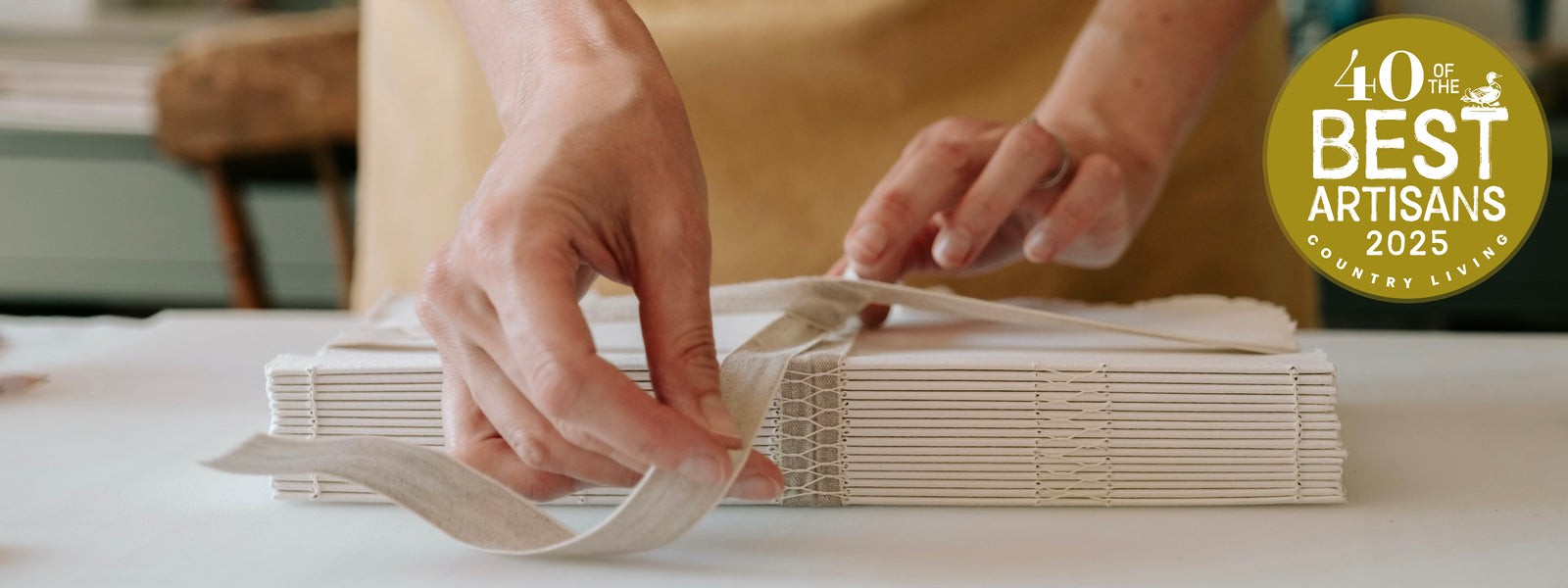 Bookbinder ties linen ribbon over cotton rag sketchbook bound with an exposed spine; the Country Living 40 best Aritsans logo appears in the top right corner. 
