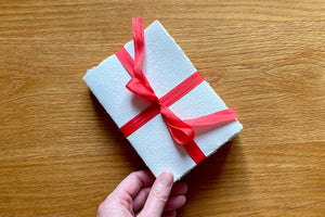 Hand holds corner of Cotton Rag Sketchbook bound at A6 portrait size with cotton rag handmade paper and red silk ribbon.