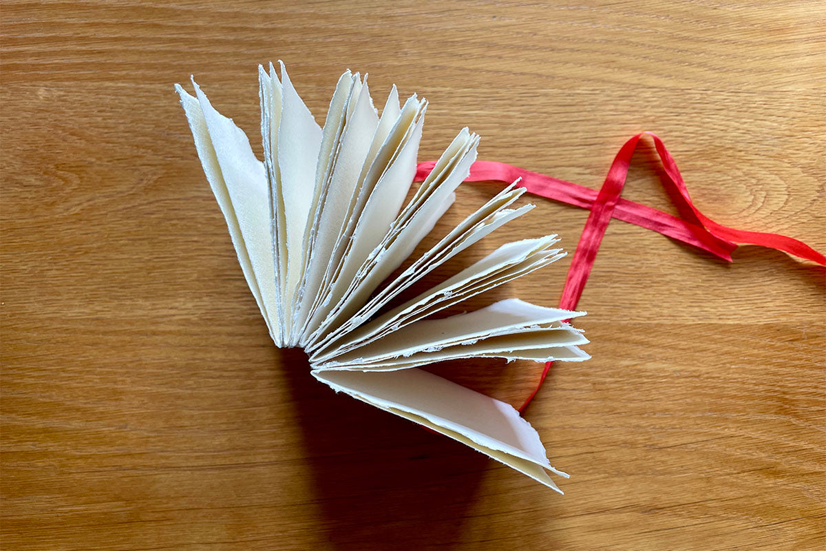 Cotton Rag Sketchbook bound with cotton rag handmade paper and tied with red silk ribbon rests on natural wood surface.