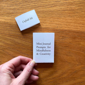 Printed titlepage of miniature artist's book reads 'Mini Journal Prompts for Mindfulness and Creativity'. Its closing belly band sits to the side, printed with 'Unfold Me'.