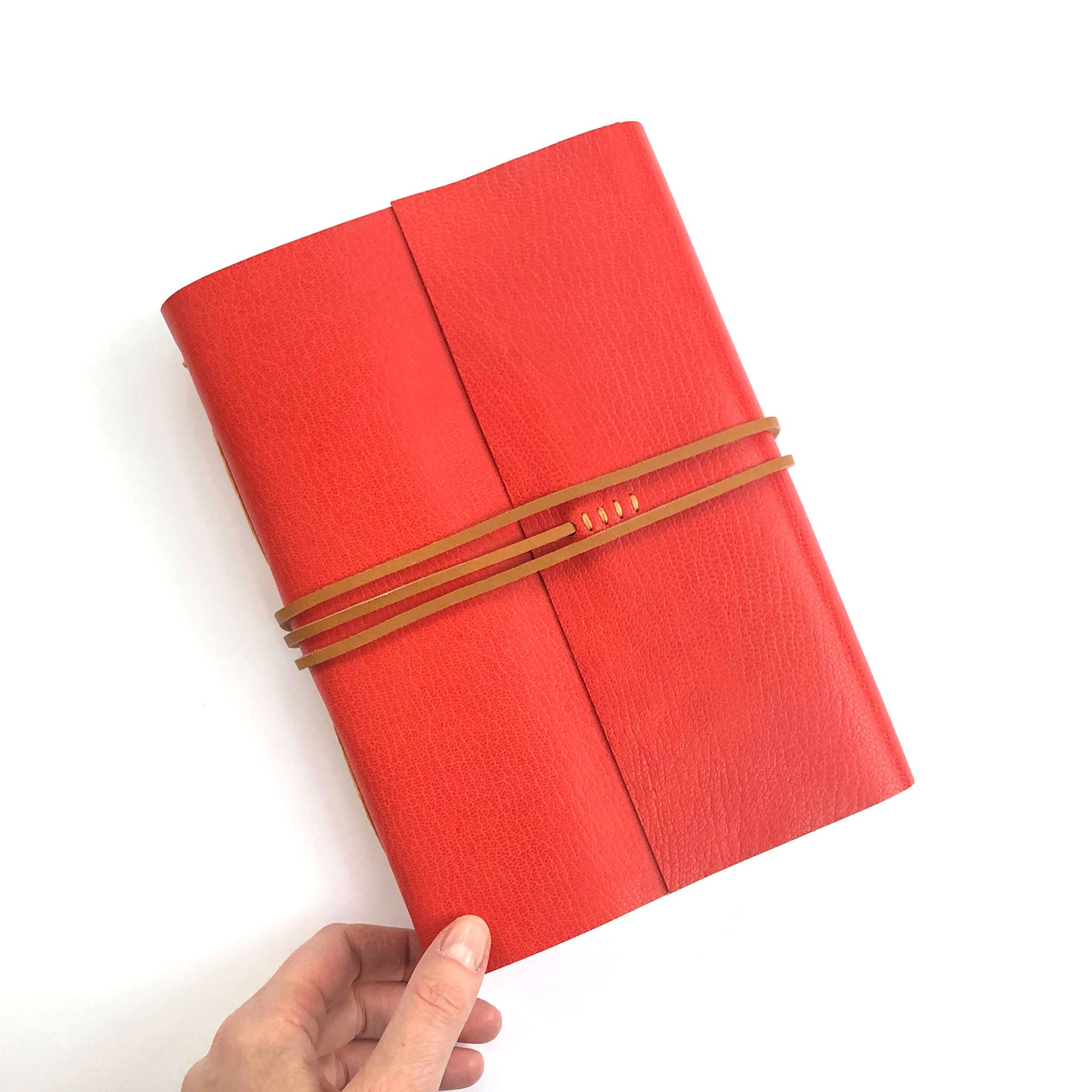 Red Leather Journal with Tan thong wrapped around the middle held at its bottom left corner by a just visible hand, against a plain white background.