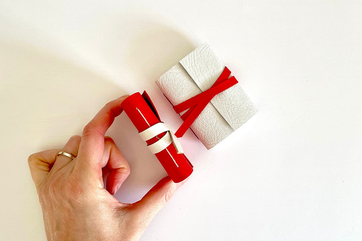 Mini Journal bound in Red leather with gift wrapping and free leporello book of writing prompts lie on a wooden surface
