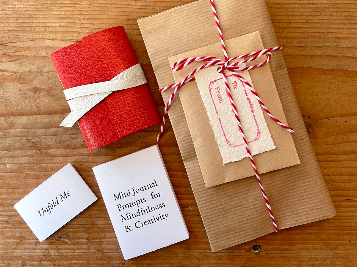 Mini Journal bound in Red leather with gift wrapping and free leporello book of writing prompts lie on a wooden surface