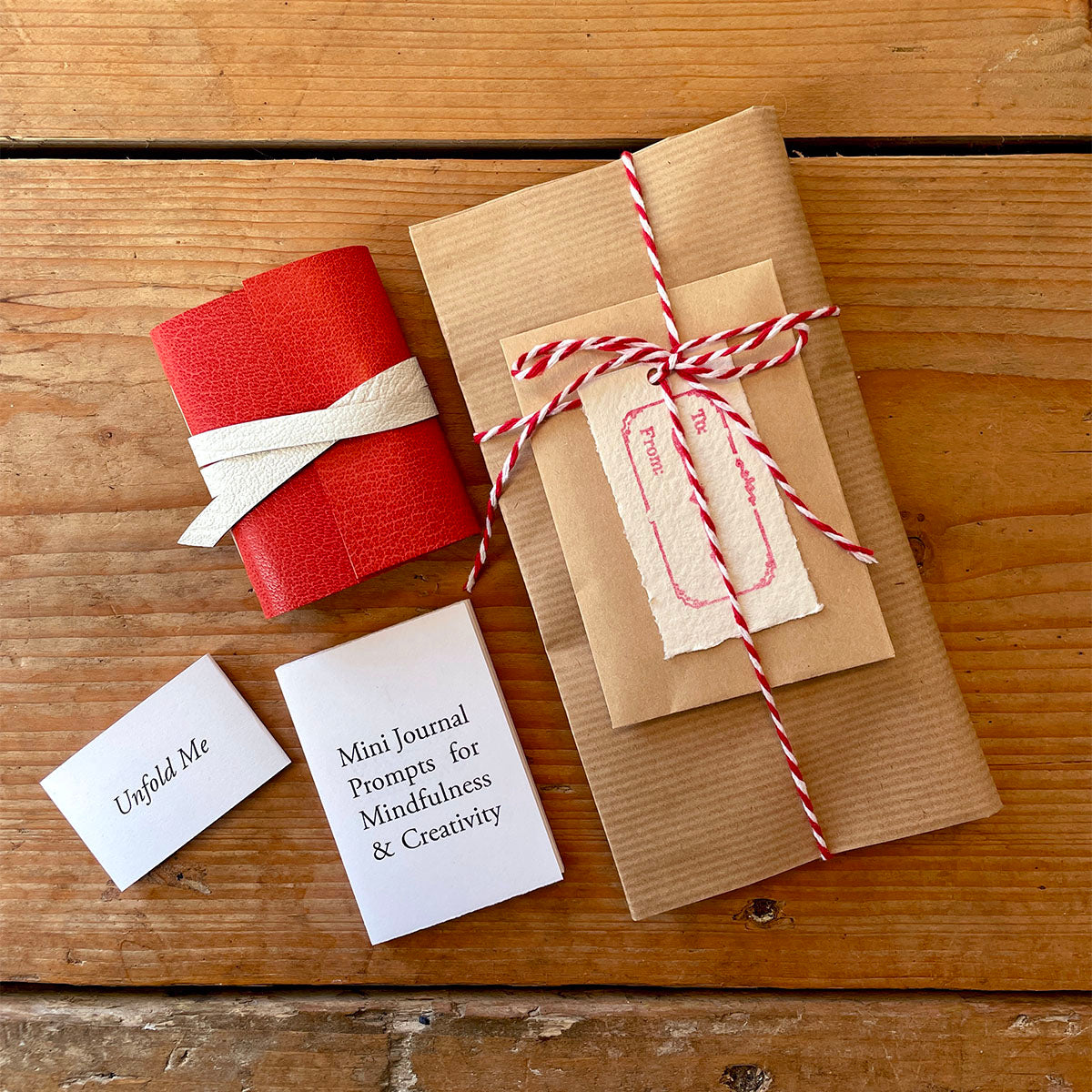 Red leather mini journal with Christmas gift wrapping sits on wooden surface with miniature leporello book of writing prompts