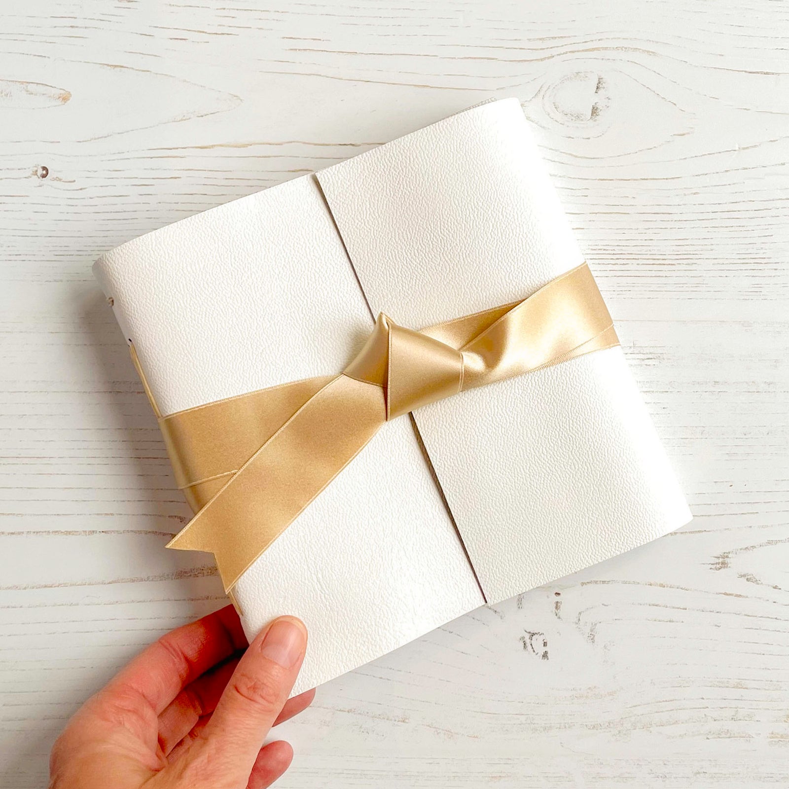 A handmade Wedding Guest Book bound in white leather with champagne silk ribbon, small square sits on a white natural wood grained surface.