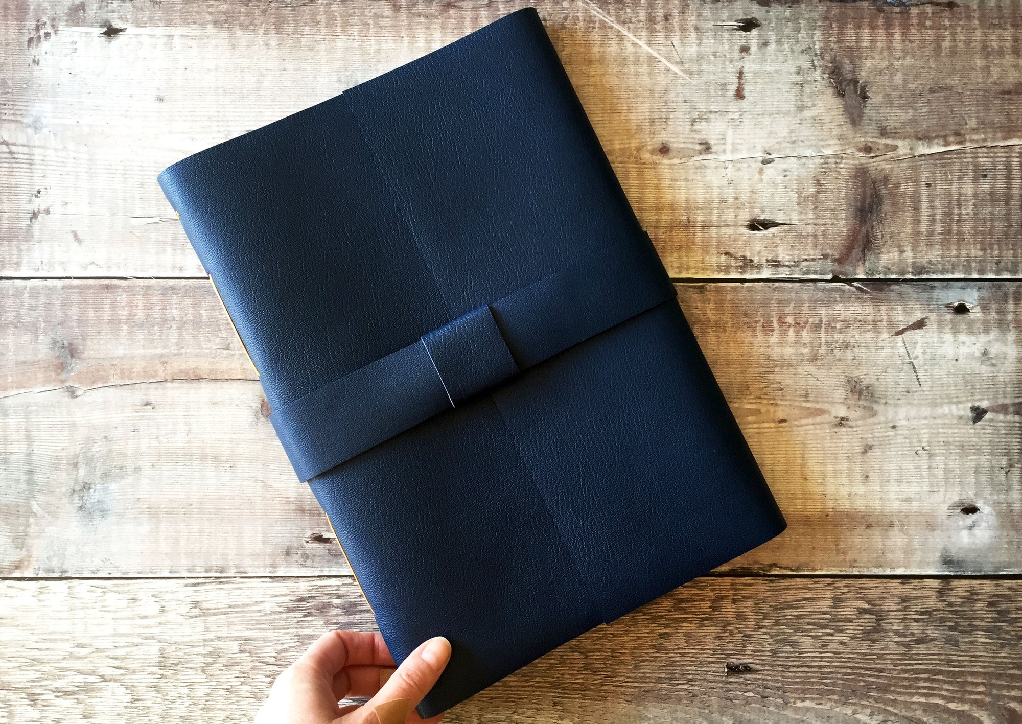 Hand holds Navy Blue A4 Leather Journal Navy Blue against wood background.